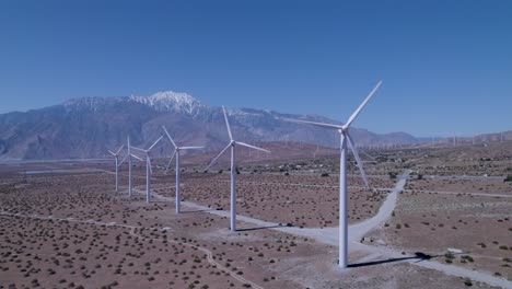 drone pulls back, revealing windmills in the desert with snow-capped mountains and additional windmills in the far background