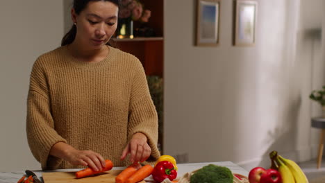 woman at home in kitchen preparing healthy fresh vegetables for vegetarian or vegan meal chopping carrots on board 1