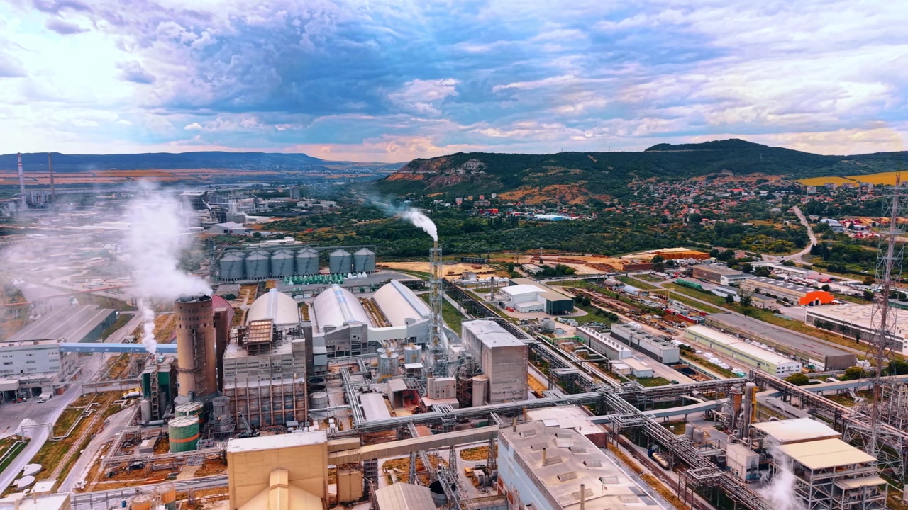 Industrial factory complex with smoking chimneys. White smoke rises from a manufacturing plant situated near a rural town and green hills under a cloudy sky.