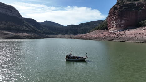 Aerial-view-of-fishing-boat-floating-in-the-low-water-of-Sau-swamp-reservoir-in-sunlit-woodland-valley,-Catalan-Spain