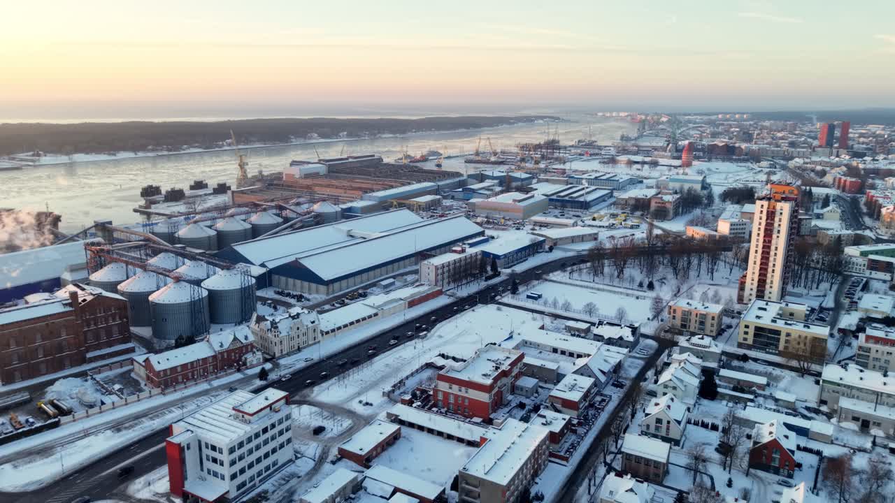 Drone-ilmakuva lumipeitteisestä kaupungista ja teollisuusrannasta, jossa on siiloja, höyrypilvi, teitä ja jäinen joki, auringonnousun aikaan kylmänä talviaamuna.