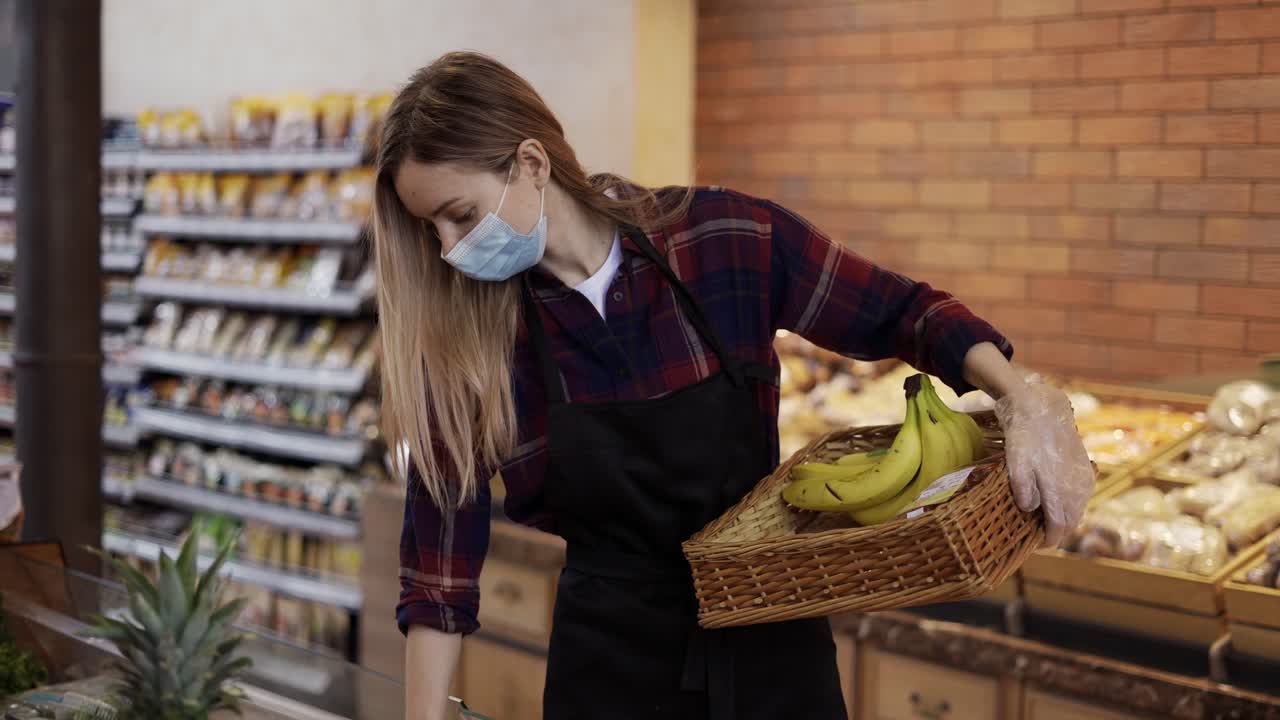 검은 아프론과 마스크를 입은 여성 노동자가 슈퍼마켓에서 과일을 다시 채우고 있습니다.