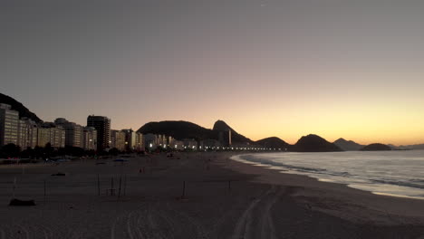 Vista-Aérea-De-La-Playa-De-Copacabana-Temprano-En-La-Mañana-Antes-Del-Amanecer-A-La-Hora-Dorada-Con-Las-Luces-De-La-Ciudad,-El-Pan-De-Azúcar-Y-Las-Colinas-De-Niteroi-Al-Fondo