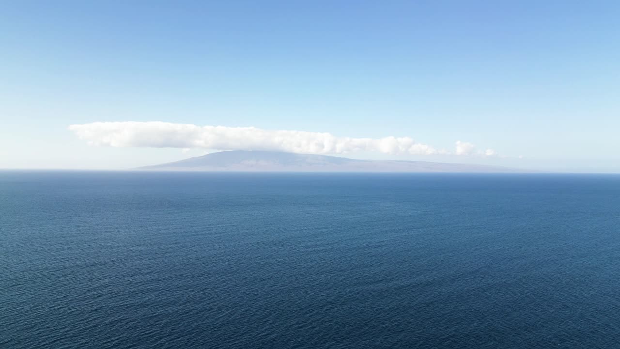 Aerial moves slowly toward a distant island beyond open water beneath blue sky