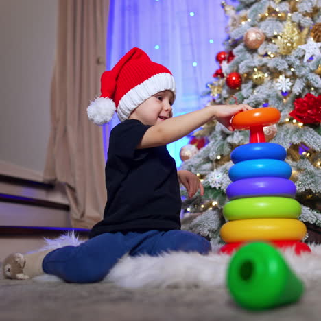 cheerful baby boy putting the pyramid rings on the basis. cute kid in santa cap playing near the christmas tree. low angle view.