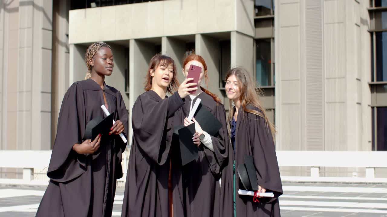 Diverse female graduates walking on plaza pulling smartphone taking selfie celebrating graduation