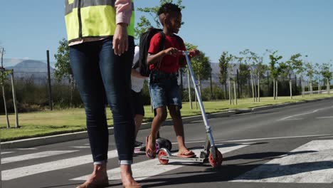 Dos-Niños-Con-Mochilas-Escolares-Montando-Scooters-Y-Cruzando-La-Calle