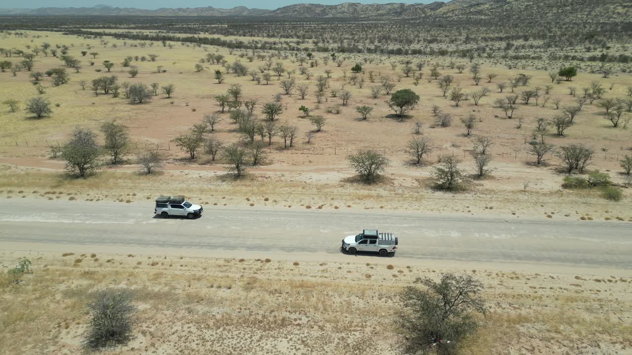Vista aerea da drone nel deserto della Namibia, con un'auto SUV che percorre una strada stretta, sabbia e alberi verdi ai lati e montagne all'orizzonte in una giornata di sole