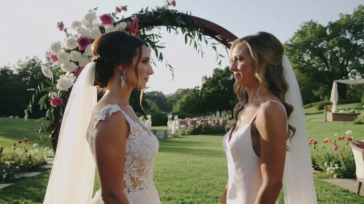 Starting right bride reciting wedding vows beneath garden floral arch, left bride listening, gowns
