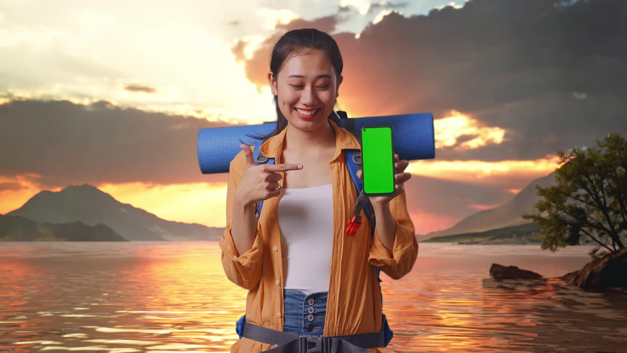 Asian Female Hiker With Mountaineering Backpack Smiling And Pointing To Mock Up Green Screen Smartphone While Standing At A Lake During Sunset Time