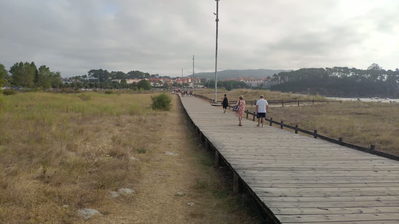an elderly couple strolls over a wooden overpass near the beach on a cloudy gray morning, taking a panoramic view. Porto-novo, Galicia, Spain