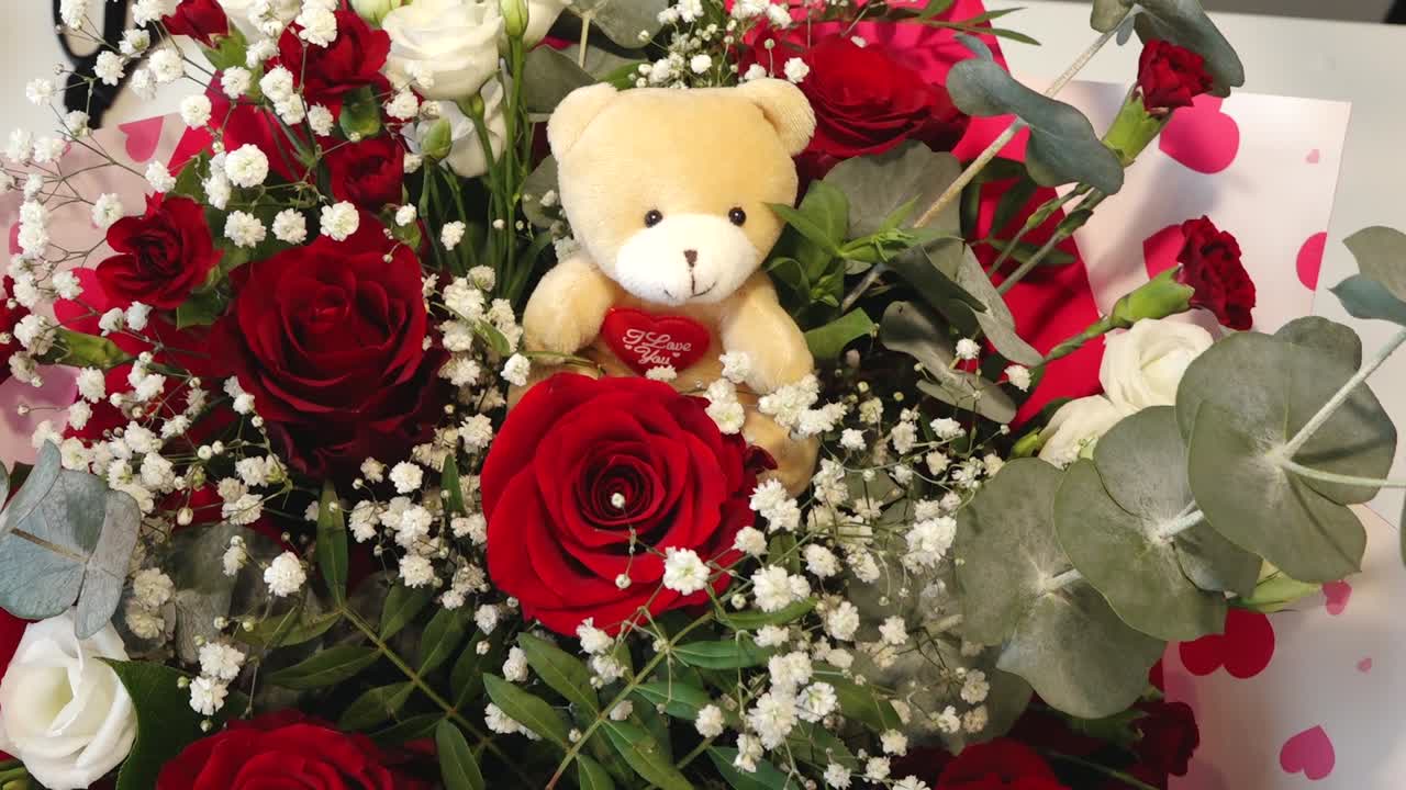 a soft teddy bear nestled among vibrant red roses, white baby's breath, and green foliage. Romantic floral arrangement with a warm, affectionate mood