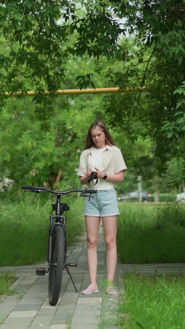 여성은 검은 장갑을 들고, 포장 된 길에서 주차 된 자전거에 접근합니다. 스마트워치가 오른쪽에 보일 때 장갑을 착용합니다. 배경에는 울창한 초록색, 나무 및 거리에서 지나가는 자동차가 포함됩니다.