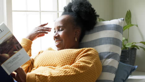 happy african american senior woman lying, reading book and smiling in sunny room, slow motion