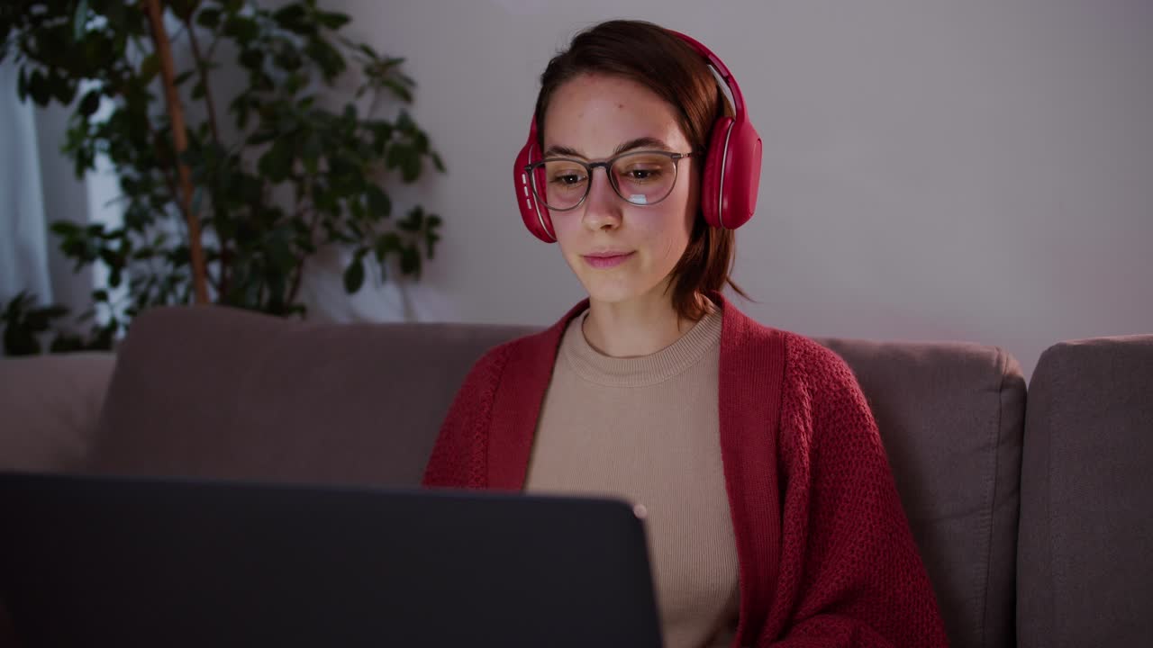 A happy and focused brunette girl in a red sweater with glasses and wireless headphones studies foreign languages using online lessons on a gray laptop sitting on the sofa in a modern apartment in the evening