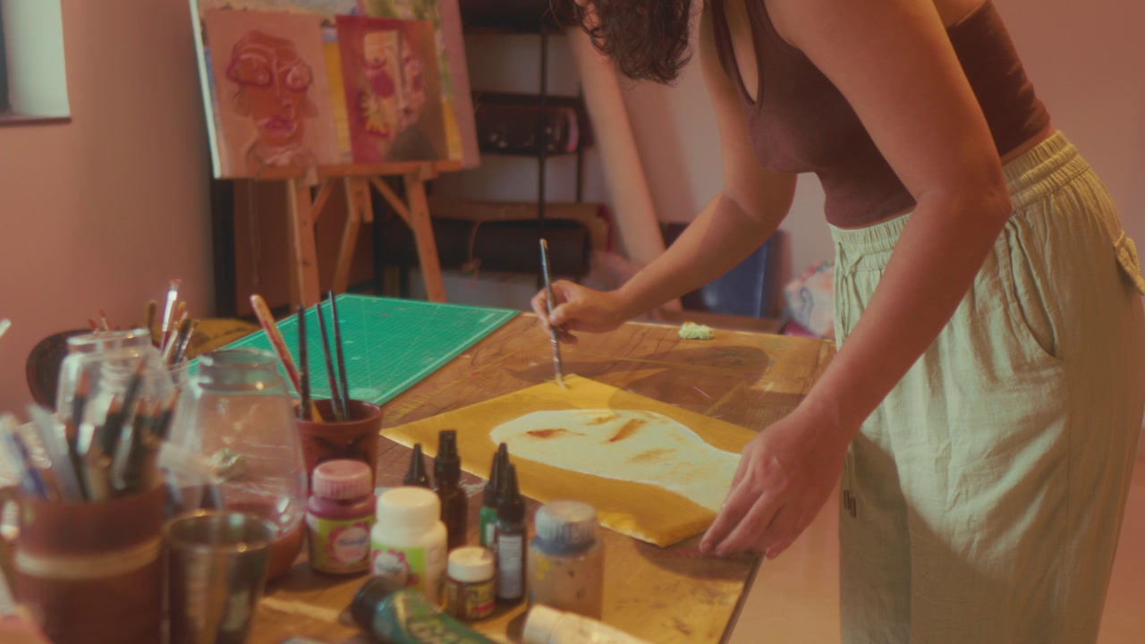 Wide shot of woman painting at desk in art studio surrounded by tools and materials. Calm creative workflow, ideal for artistic lifestyle, hobby and design process themes.