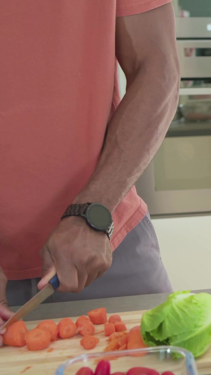 Prendendo il coltello da chef, un uomo con una maglietta color corallo affetta carote su un tagliere in cucina, disponendo i pomodori