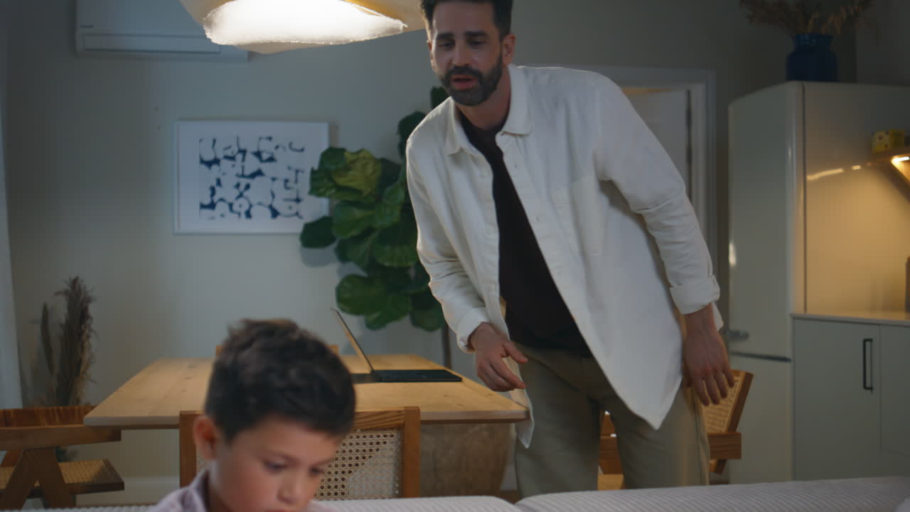 Bearded man working laptop at table boy with smartphone resting couch in home