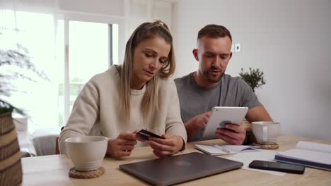 caucasian couple sitting at kitchen table online shopping using digital tablet