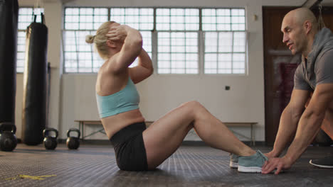 Fit-caucasian-woman-performing-crunches-while-male-trainer-holding-her-legs-at-the-gym