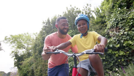 happy african american father teaching son to ride bicycle in sunny garden, slow motion