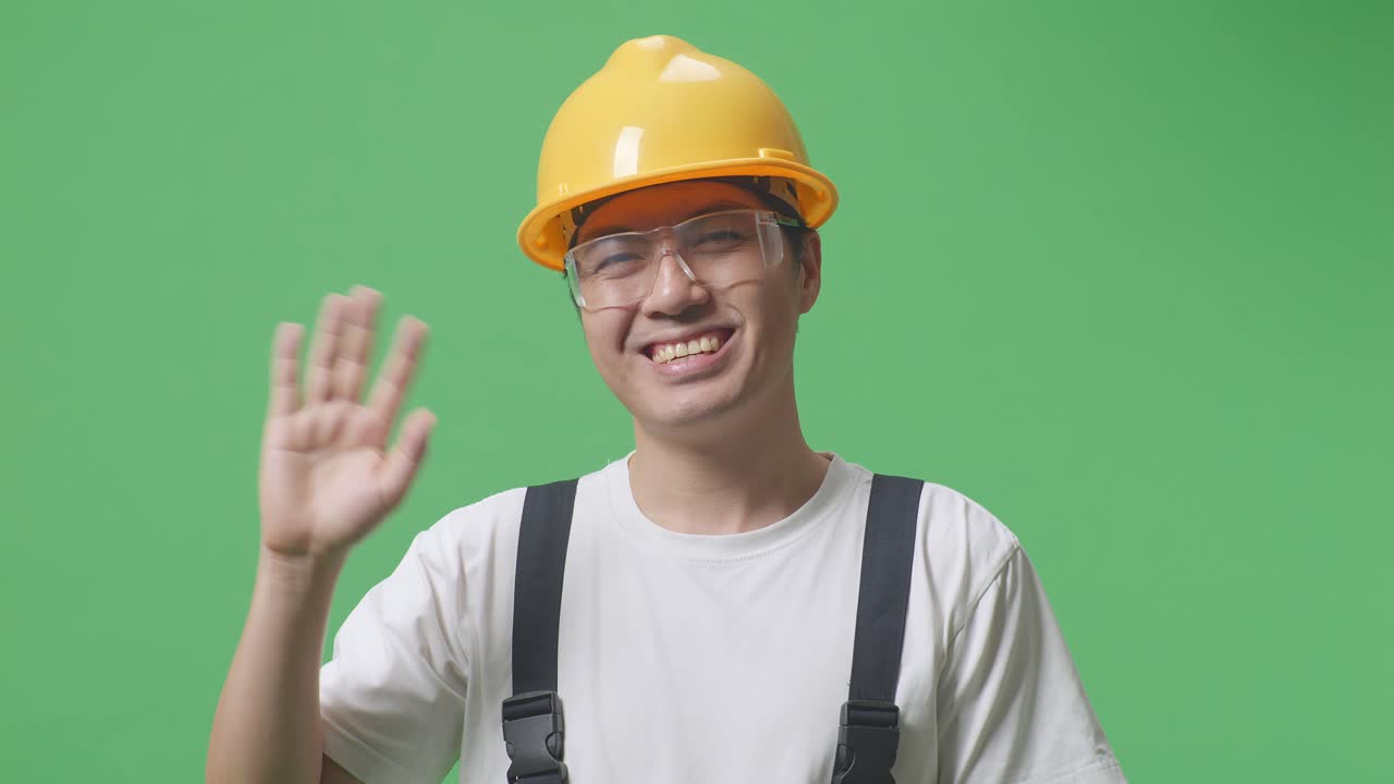 안경과 안전 헬을 착용한 아시아인 노동자가 미소 짓고 손을 흔들며 녹색 스크린 배경 스튜디오에 서서 작별 인사를 하고 있습니다.