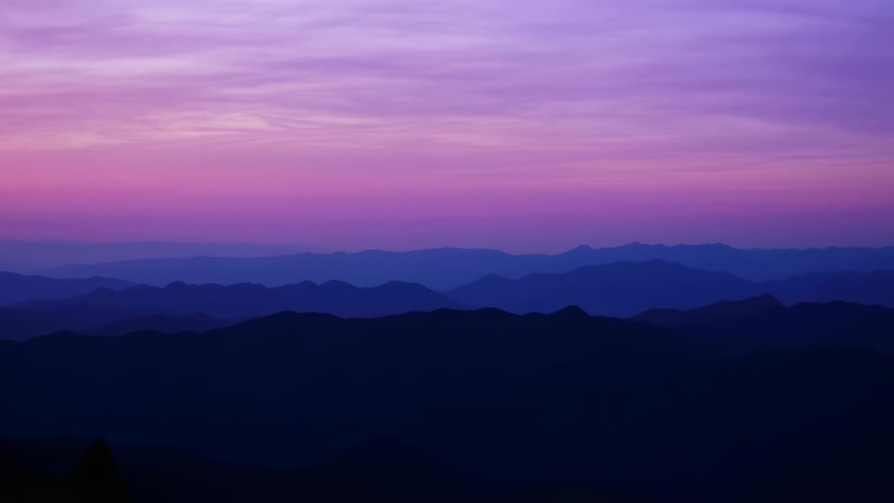 Creste montuose scure sovrapposte si allontanano verso l'orizzonte sotto un cielo pastello al tramonto. Colori sfumati delicati passano dal viola intenso al rosa e all'arancione pallido