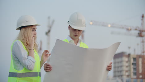 two builders with drawings standing on the background of buildings under construction in helmets and vests a woman talking on the phone with the customer.