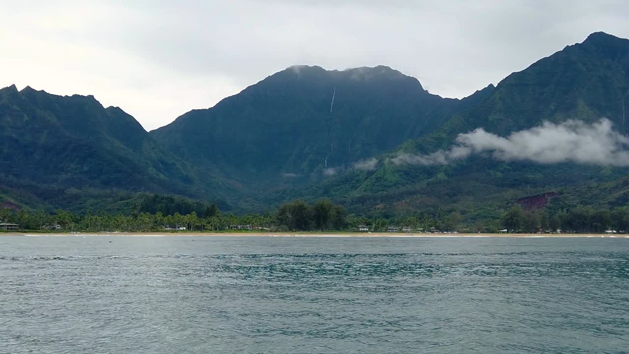 hd 120fps 하와이 카우아이는 오른쪽에서 왼쪽으로 떠다니는 바다 위에 해변과 구름이 있는 계곡에 구름이 있는 산과 흐린 거리에 폭포가 있고 약간 오른쪽에서 왼쪽 팬으로 보트를 타고 있습니다.