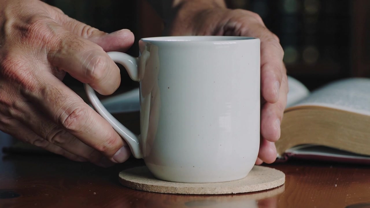 Mani con polsini blu che abbassano e posizionano una tazza da sopra un libro aperto su un sottobicchiere nello studio di casa, proteggendo il libro.