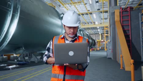 ingeniero masculino asiático con casco de seguridad trabajando en una computadora portátil mientras está de pie en una fábrica de fabricación de tuberías