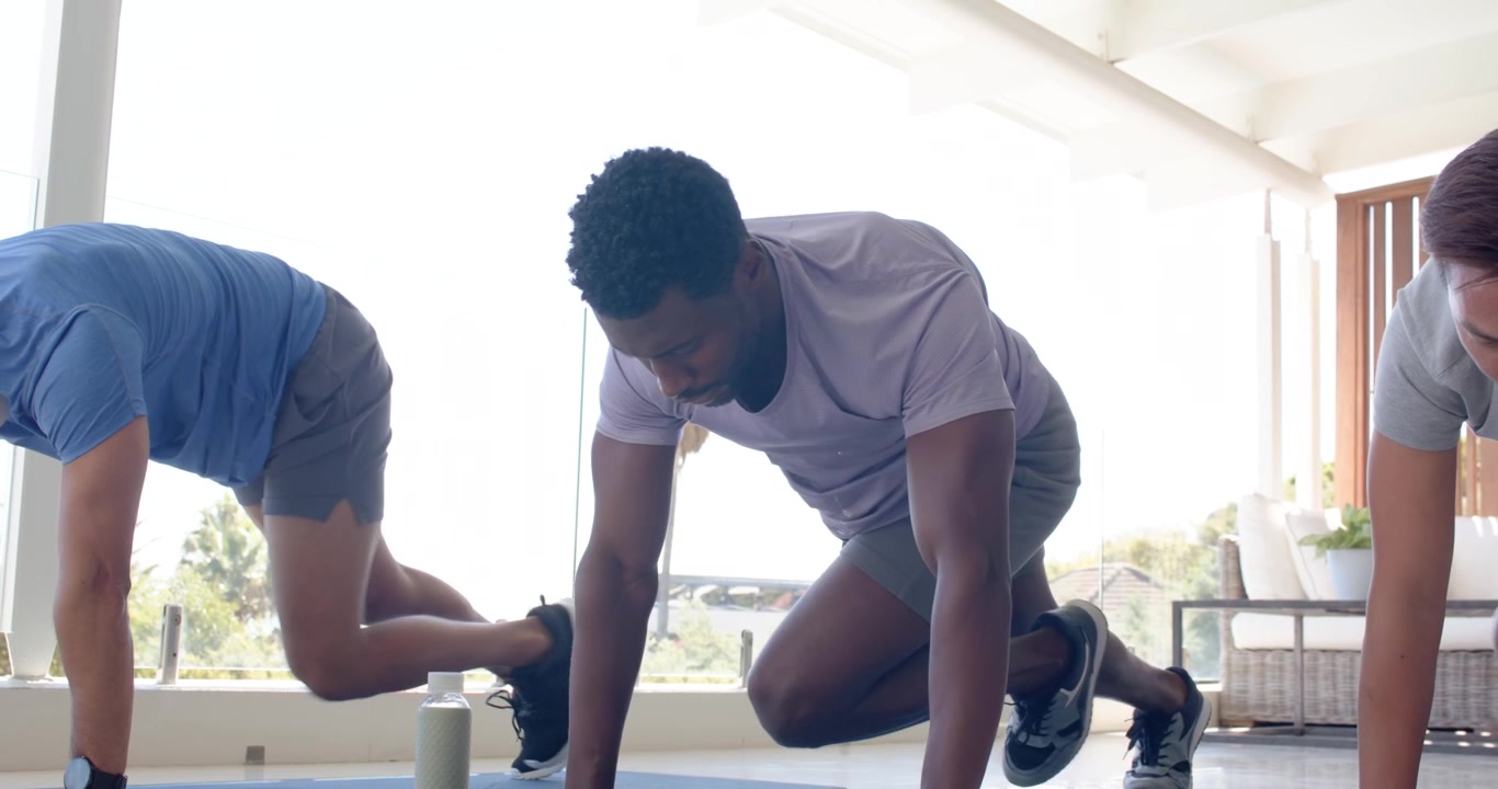 Diverse male trio starting plank, driving alternating knee tucks on blue mats on patio with bottle