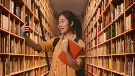 side view of asian woman student with a backpack and some books having a video call on smartphone while standing in the library