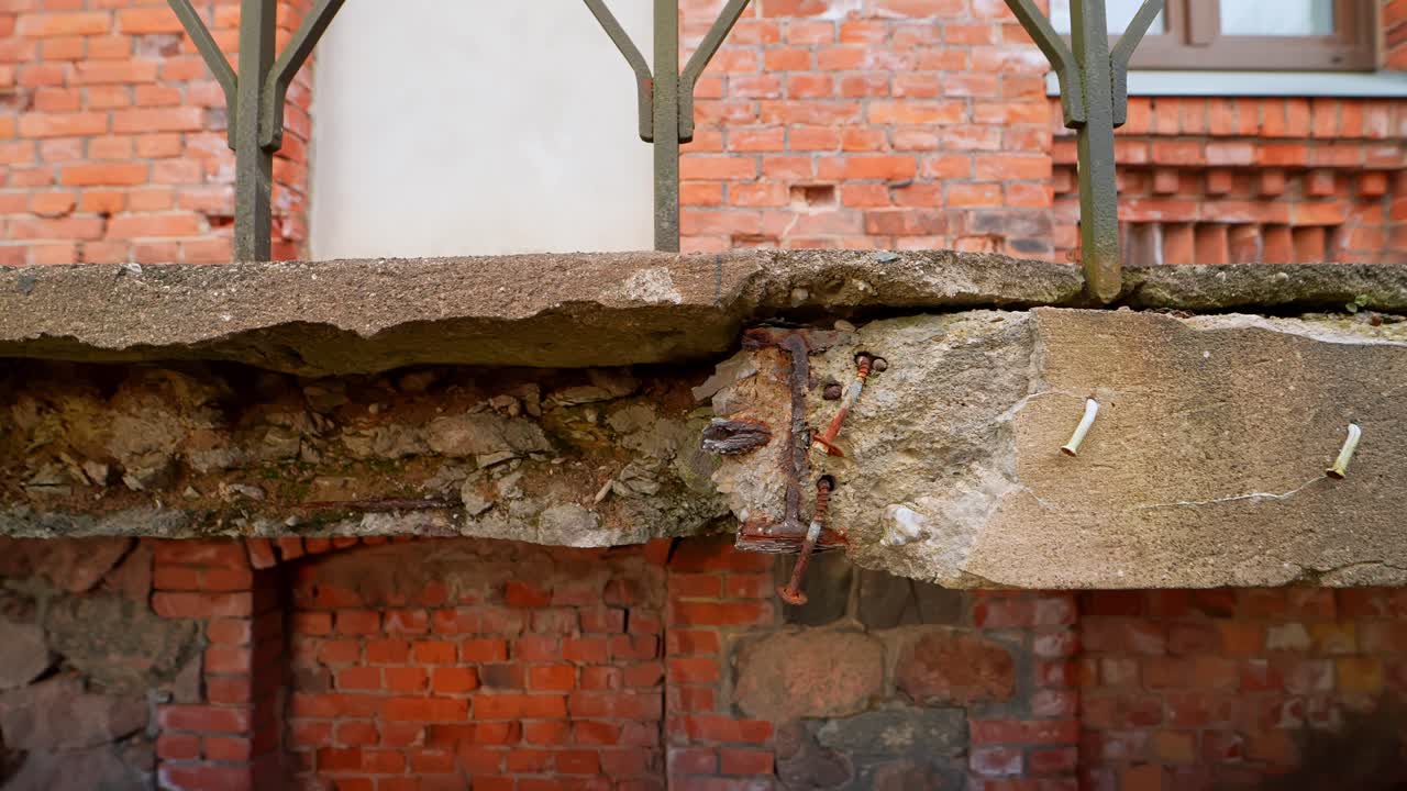 Primo piano di una lastra di balcone fatiscente con armatura esposta e rattoppi di cemento sopra un muro di mattoni, che mostra scarsa fattura, corrosione e deterioramento strutturale