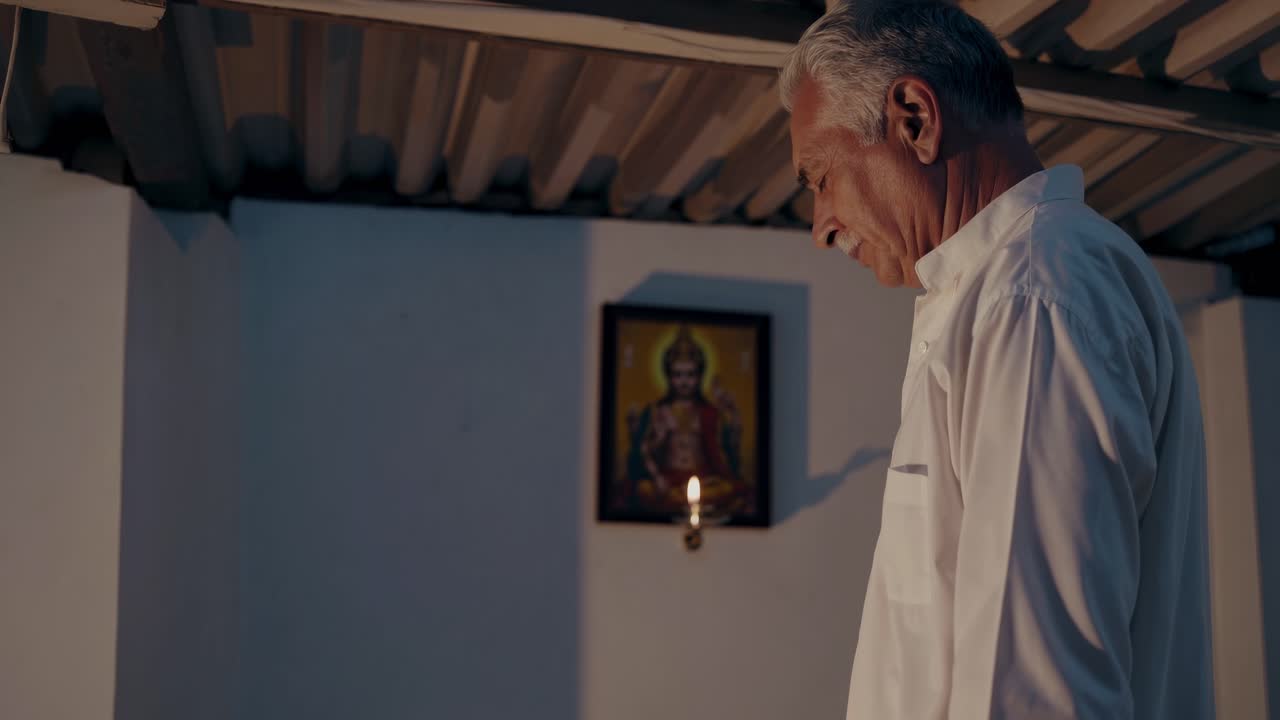 Elderly man in white shirt gazes thoughtfully at a candle-lit altar with a religious painting, reflecting on memories in a serene indoor setting