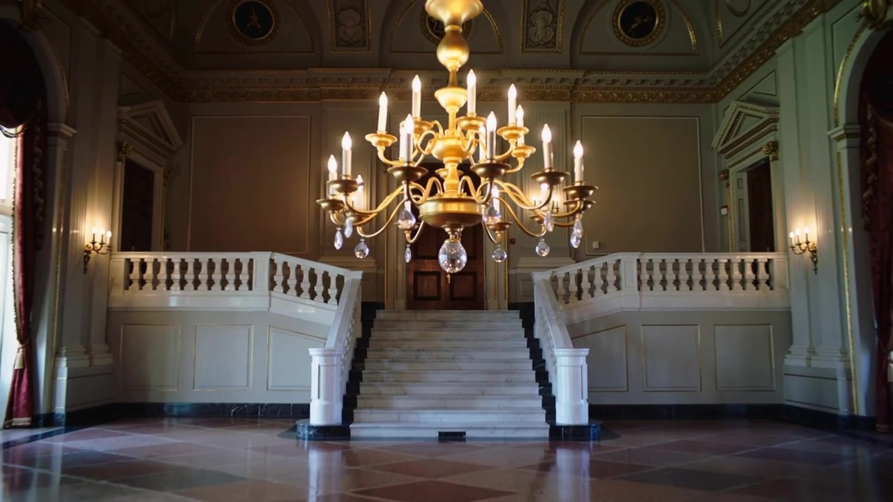 La cámara se aleja revelando un candelabro de latón y dos escaleras gemelas en un palacio, provocado por un primer plano.