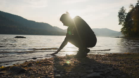 woman touches surface of water at sunset. lady leads hand on water in warm golden rays of sun in autumn evening. relaxation moment on riverbank and waiting for love