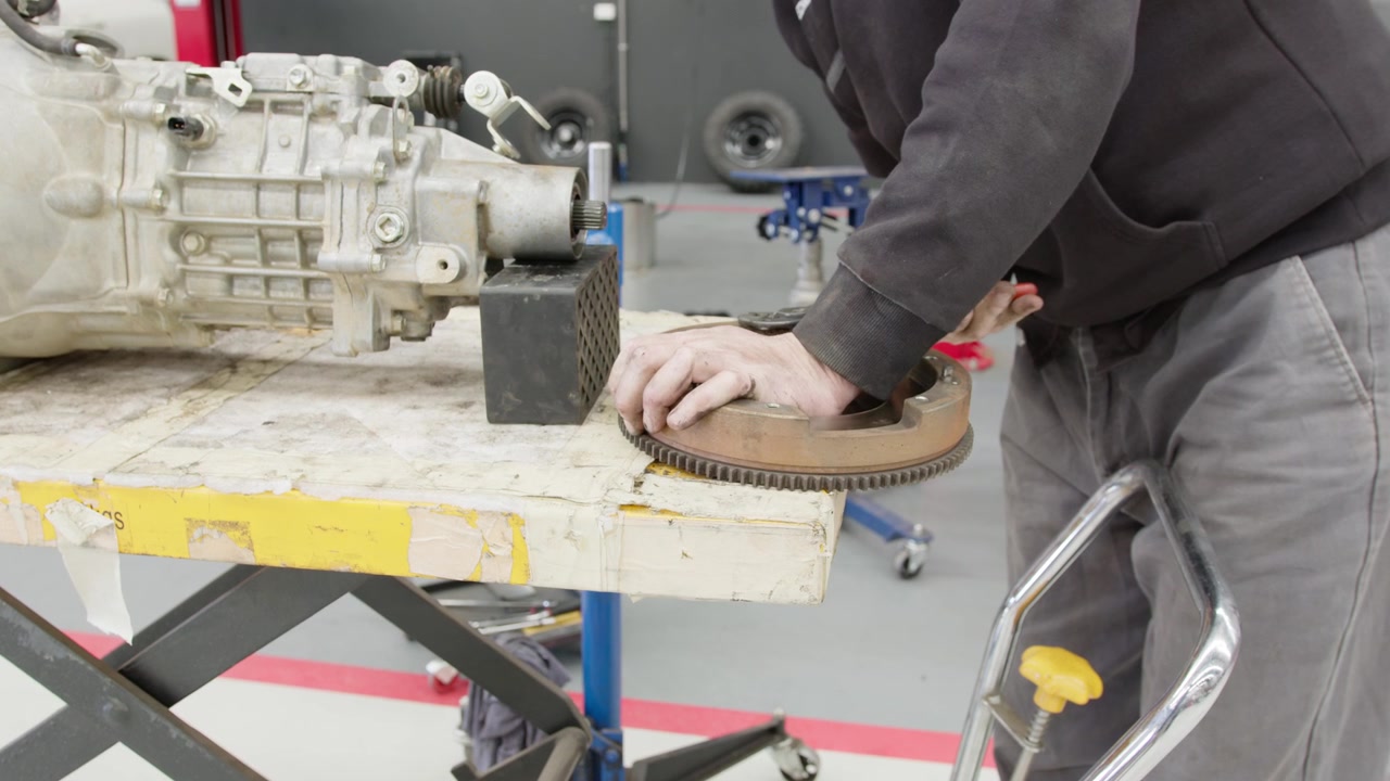 Nærbillede af en mekaniker, der inspicerer og arbejder på et svinghjul ved en værkstedsbænk. Præcis bilreparation og vedligeholdelse med professionelt værktøj i et industrielt miljø.