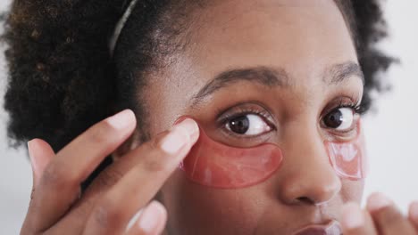 Close-up-of-african-american-woman-applying-under-eye-masks-looking-in-bathroom-mirror,-slow-motion