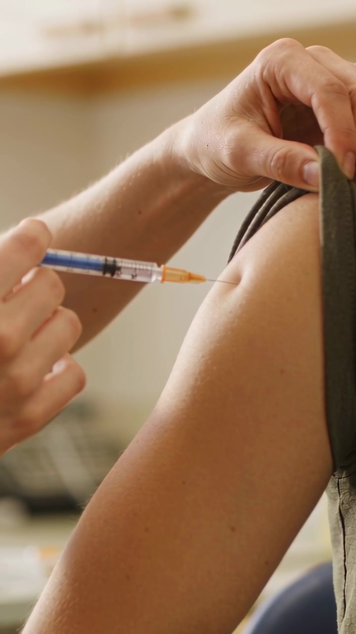 Vertical video: Pulling strap aside clinic nurse inserting orange hub syringe into arm giving dose
