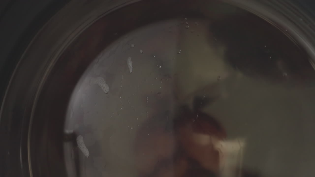 Downward view shows machine indicator above stainless panel while fabric spins inside washing machine drum, soapy water clings to glass, bubbles streak surface, modern laundromat vibe