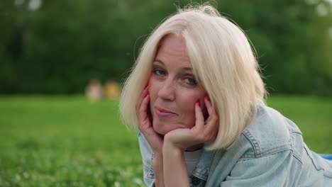 picnic mom lying on green grass with hands under chin, smiling gently, soft background of blurred park trees, casual denim jacket, warm light creating relaxed family atmosphere