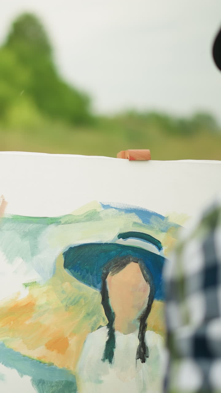 모자와 체크색 셔츠를 입은 화가가 팔레트를 들고 캔버스를 칠하고 있습니다. 배경에는 잔디 에 의자에 앉아있는 흐릿한 여성이 장면에 깊이를 더합니다.