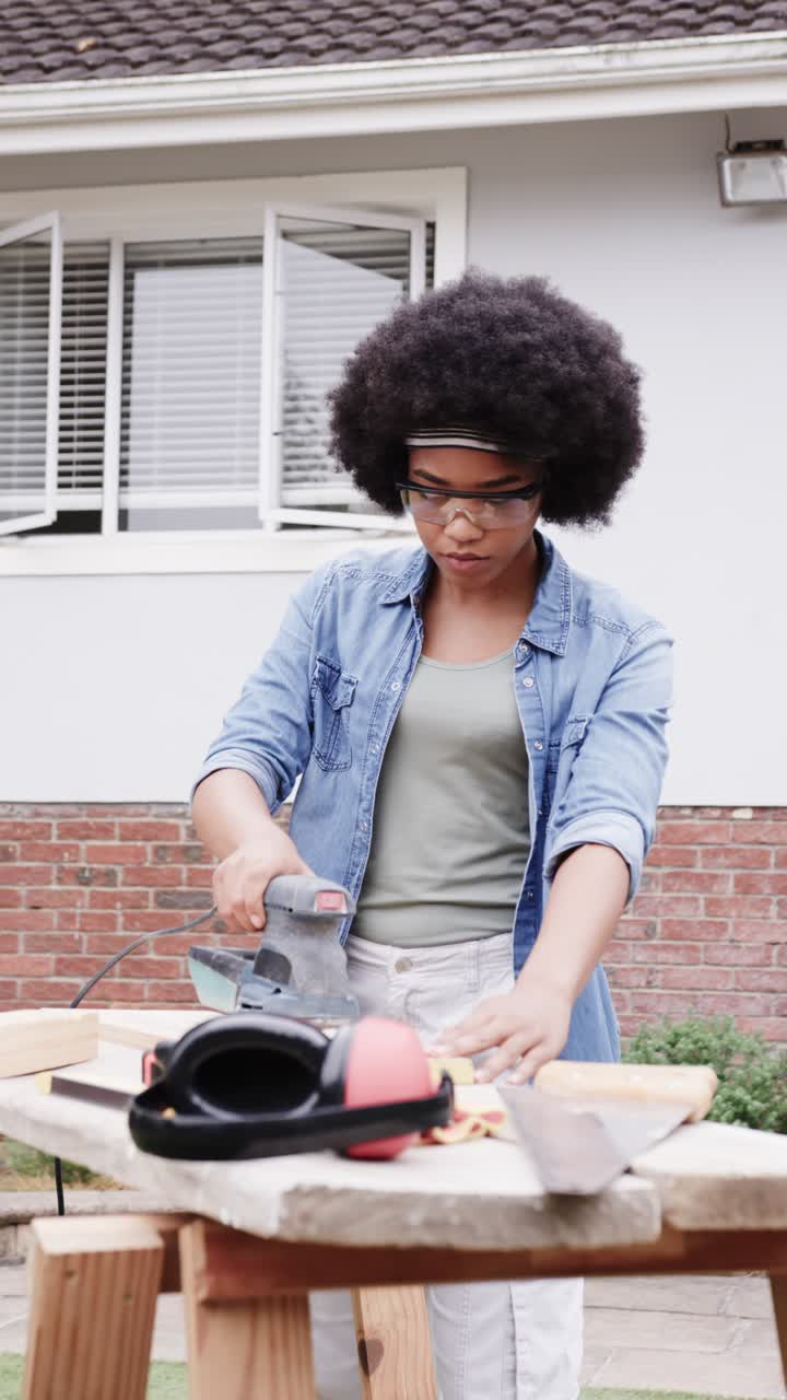 Vertical video: Placing sander woman wearing goggles, denim shirt sanding plank by house for repair