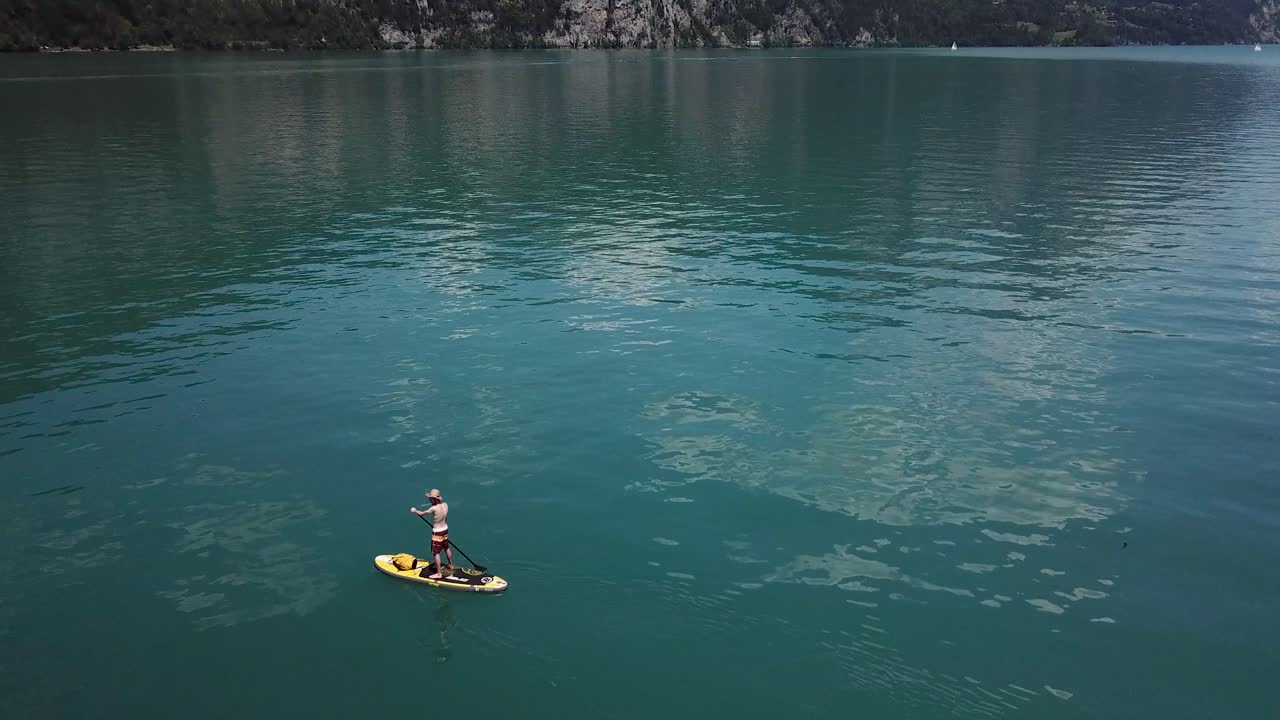 Drone, aerial orbiting shot around a male stand up paddler on a yellow sup, stand up paddle, he wears a straw hat in the middle of a lake in Switzerland