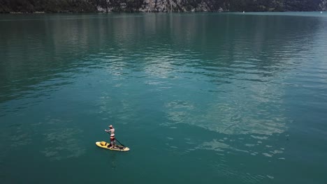 Dron,-Toma-Aérea-En-órbita-Alrededor-De-Un-Remero-De-Pie-Masculino-En-Un-Sup-Amarillo,-Remo-De-Pie,-Lleva-Un-Sombrero-De-Paja-En-Medio-De-Un-Lago-En-Suiza