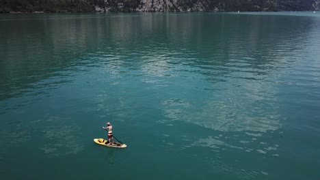 dron, toma aérea en órbita alrededor de un remero de pie masculino en un sup amarillo, remo de pie, lleva un sombrero de paja en medio de un lago en suiza