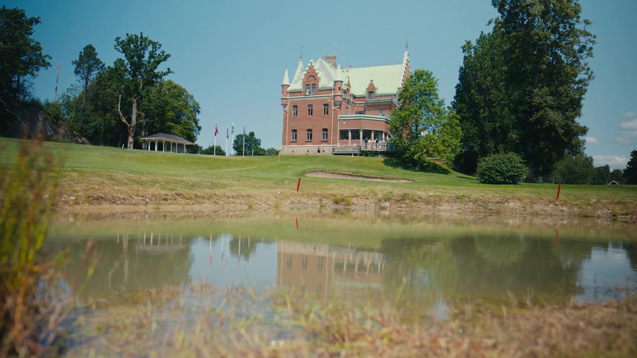 Um castelo histórico ergue-se além de um gramado bem cuidado e águas calmas, com sua fachada suavemente refletida na superfície abaixo, criando uma elegante cena de verão de patrimônio, paisagem e arquitetura.