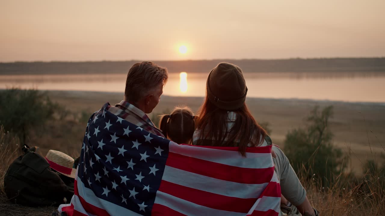 una familia feliz envuelta en la bandera de los estados unidos de américa se sienta en una alfombra y observa una hermosa puesta de sol vespertina en el verano durante su picnic fuera de la ciudad