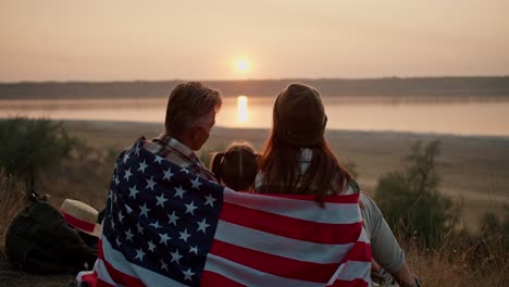 A-happy-family-wrapped-in-the-flag-of-the-United-States-of-America-sits-on-a-mat-and-watches-a-beautiful-evening-sunset-in-the-summer-during-their-picnic-outside-the-city